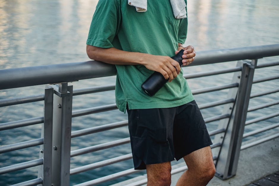 A person wearing a green t-shirt and black shorts is standing along a metal railing on a waterfront promenade, holding a black water bottle in their hands. The background features calm water with subt