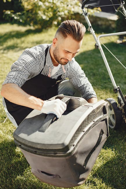 A man with styled dark hair and a beard, wearing a black apron and white gloves, is kneeling on a lush green lawn as he performs garden maintenance with a leaf blower, which is positioned on the groun